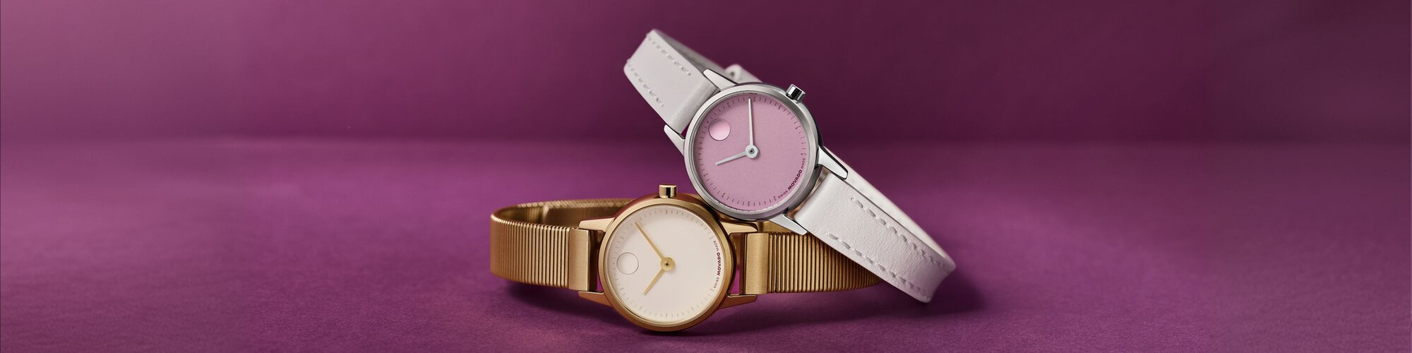 Baby Face watch with grey strap and purple dial leaning on a gold tone Baby Face watch with mesh bracelet and white dial.