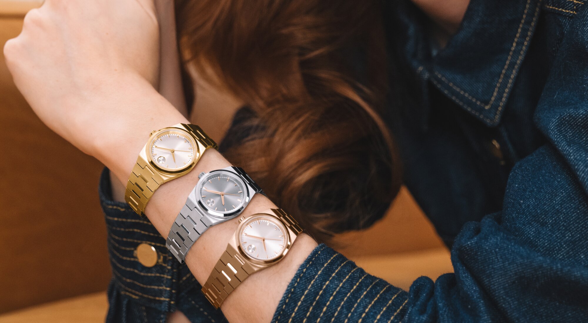 Woman in a denim shirt wearing three BOLD Quest Mini watches starting with a gold tone watch, followed by a silver watch and ending with a rose gold tone watch.