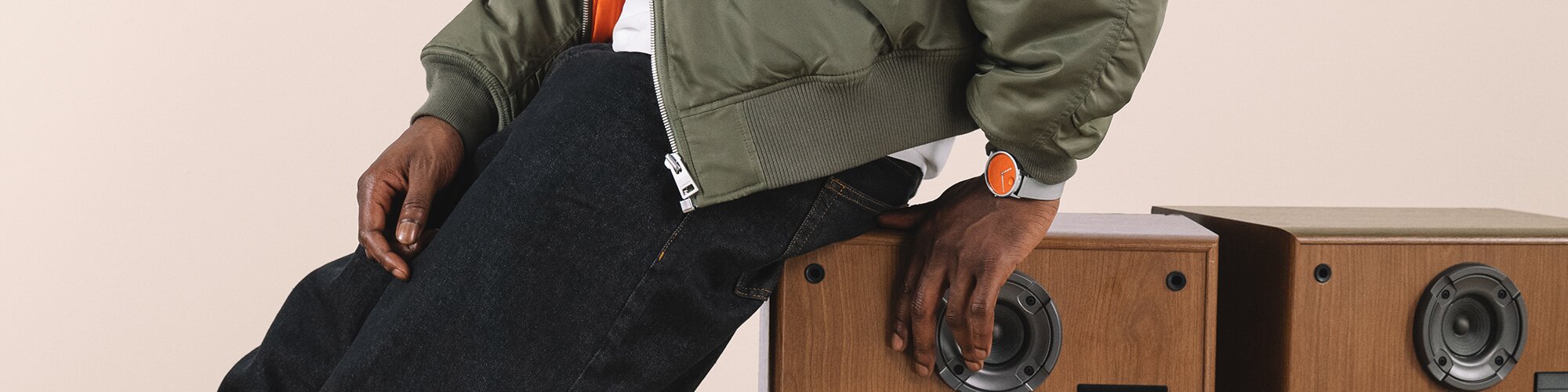 Man in a green jacket, leaning on a speaker, wearing a Movado Face watch with grey strap and orange dial 