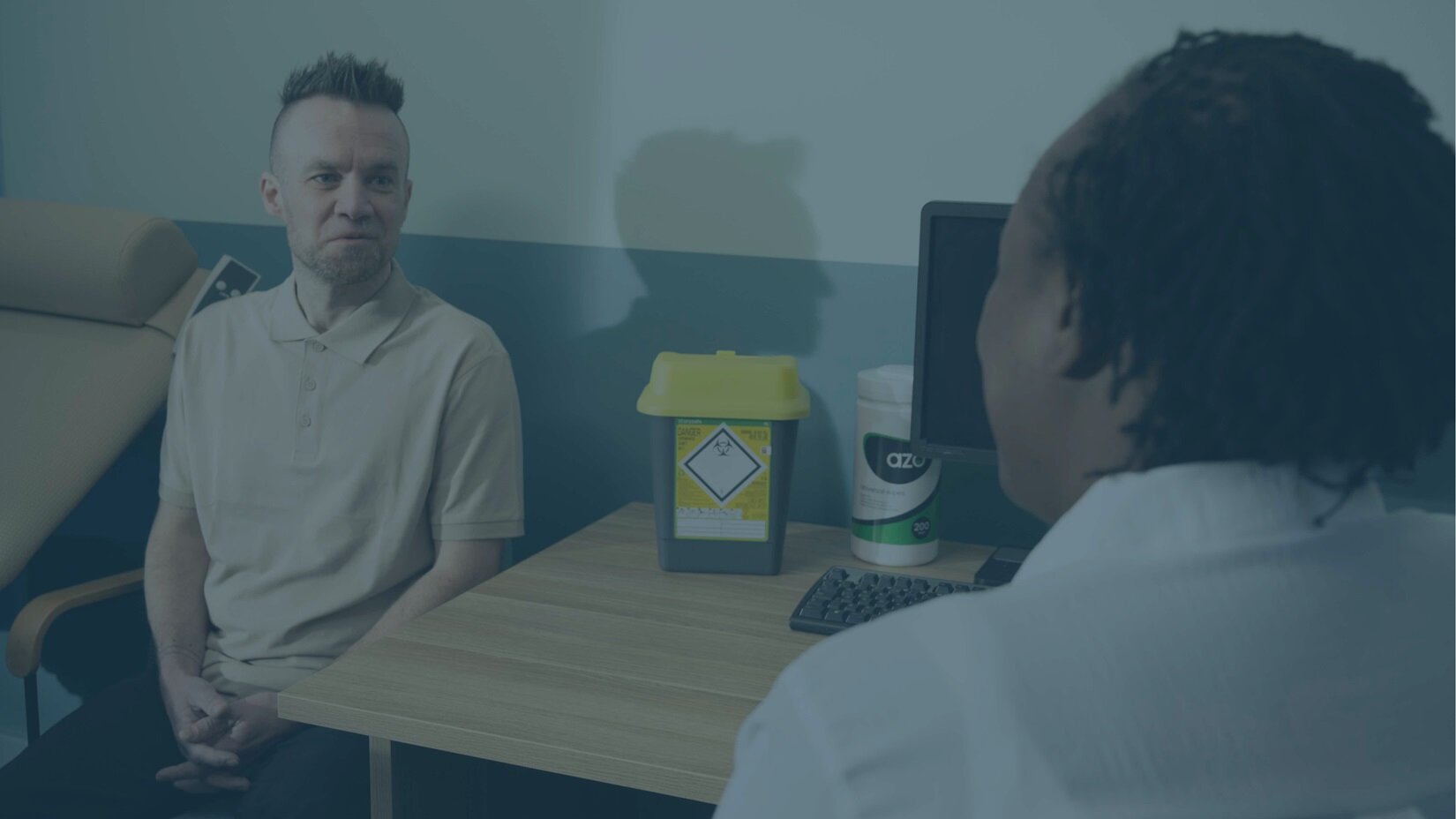 A patient sits on one side of a desk, facing a medical professional. A sharps container is prominently displayed between them.