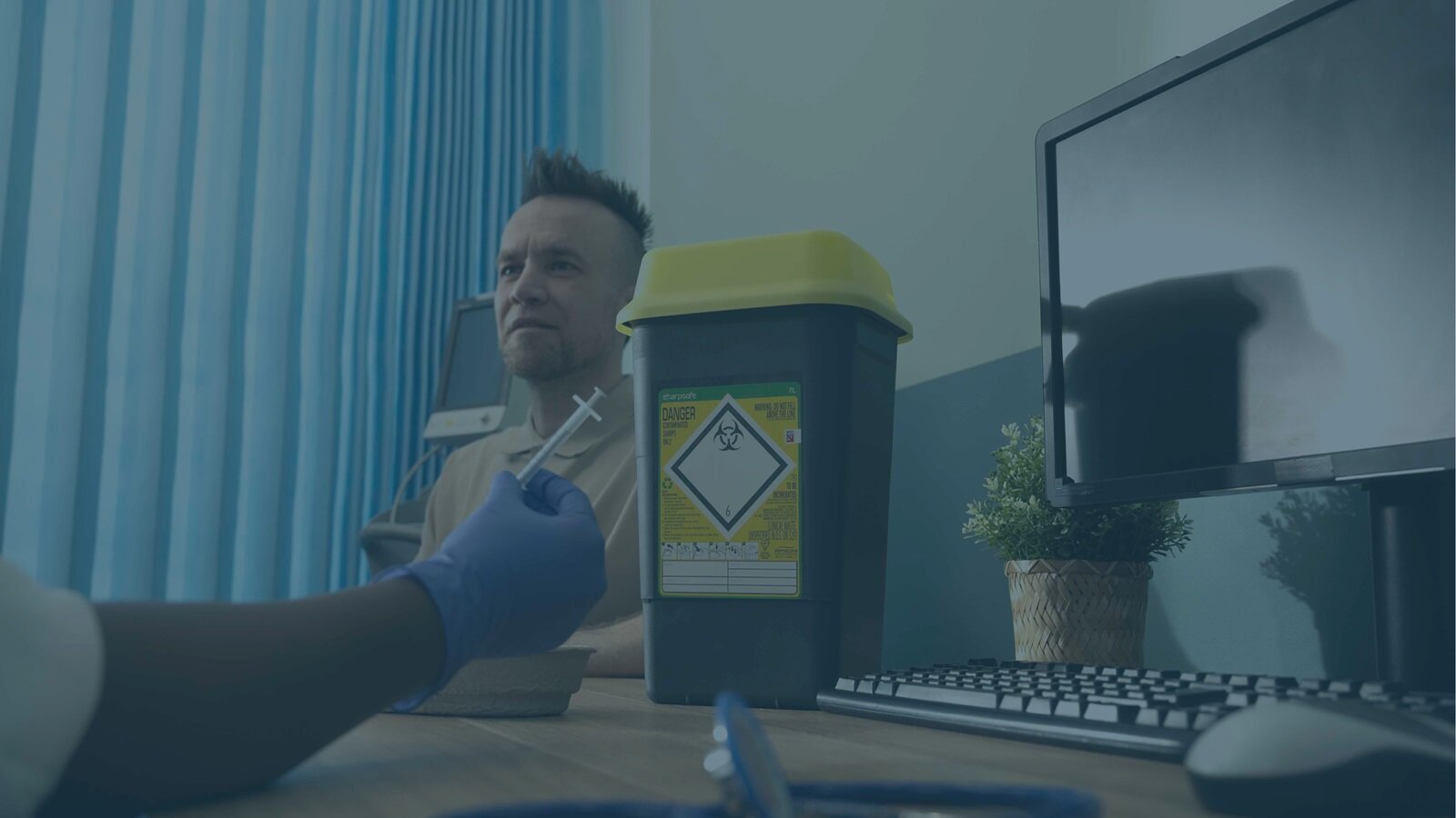 A new 7L Sharpsafe container on a GPs desk whilst she is holding a needle talking to a patient