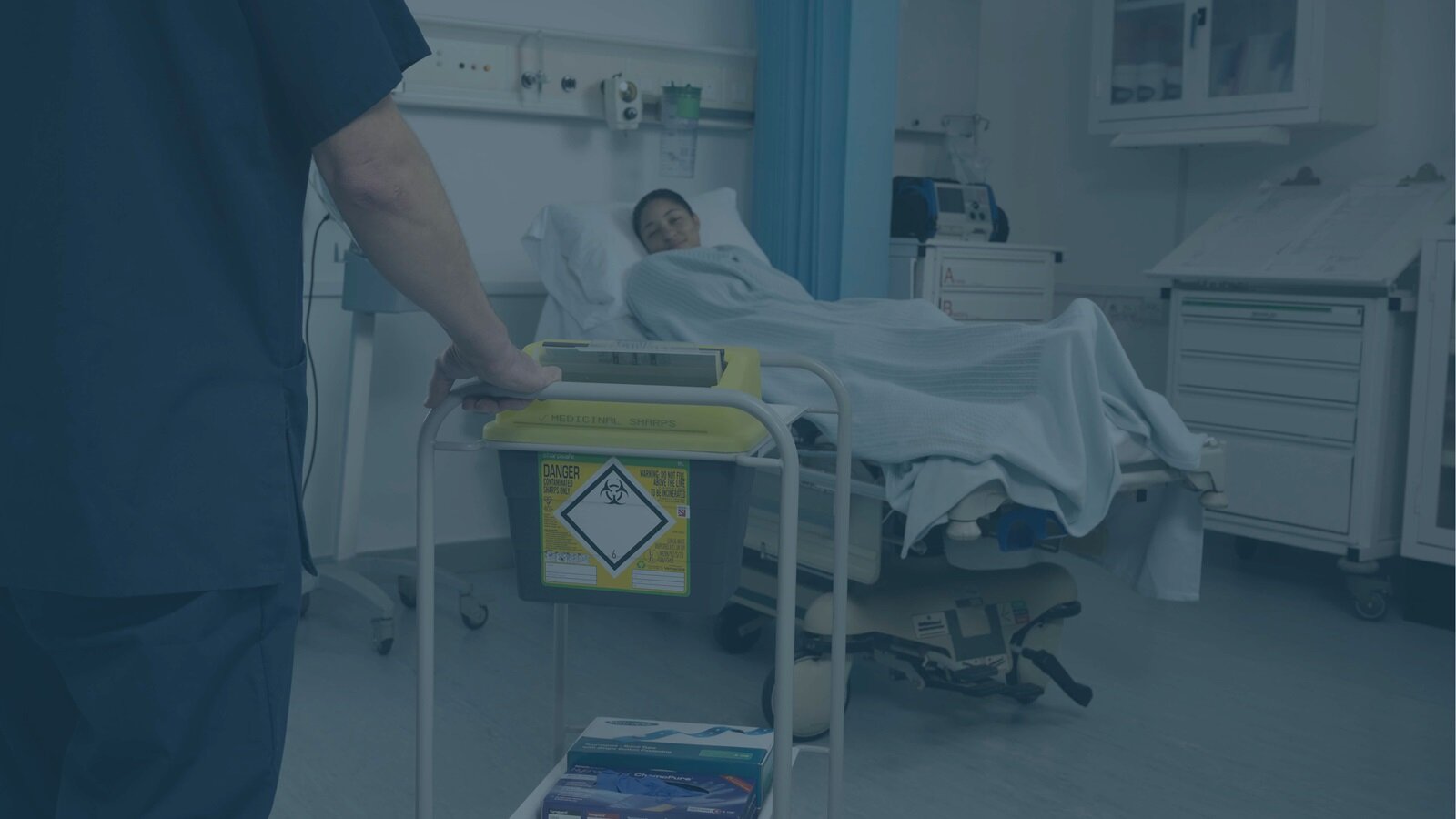 Image of a woman reclining in a hospital bed, with a Sharpsafe sharps container being wheeled towards her on a trolley.