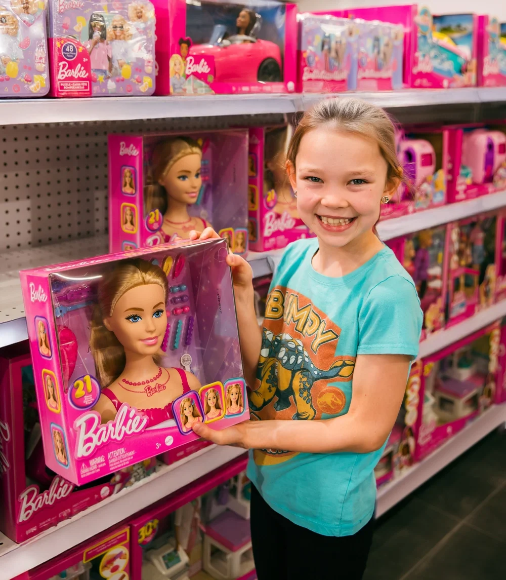 A smiling girl holding a Barbie styling head set in front of a bright pink Barbie display at Go! Toys and Games