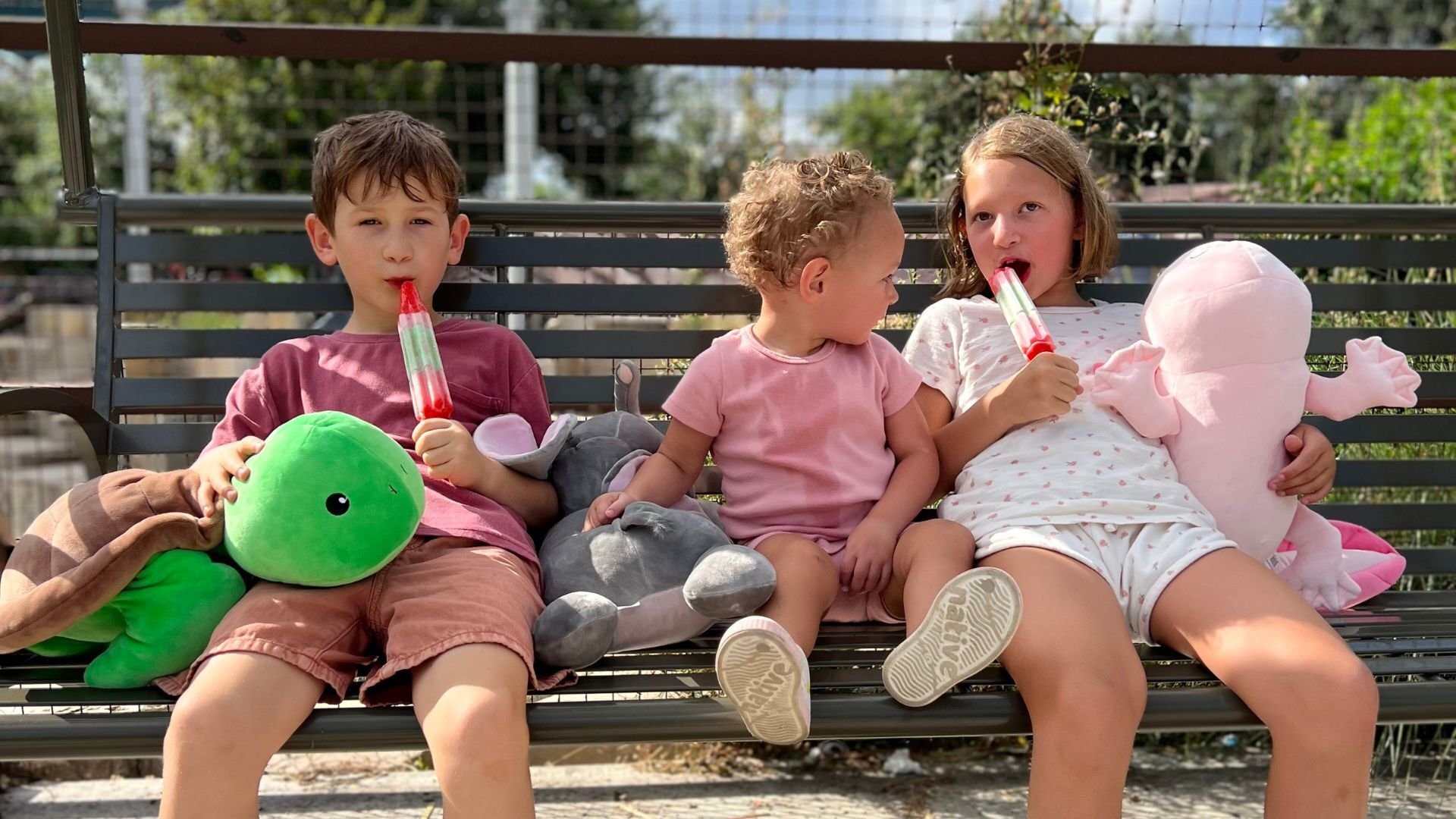 Three kids sitting on a bench eating popsicles with their Snoozimal plush friends on a sunny day