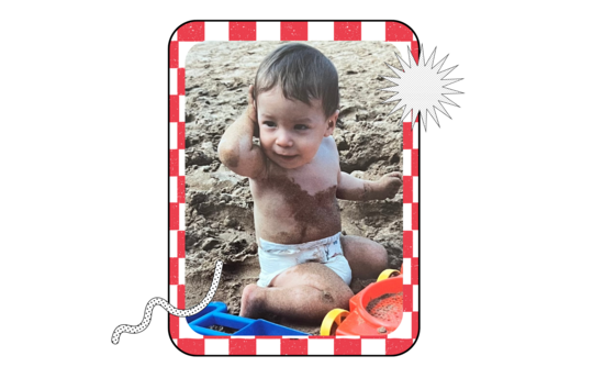 Young child sitting in the sand wearing a diaper, smiling and playing with beach toys