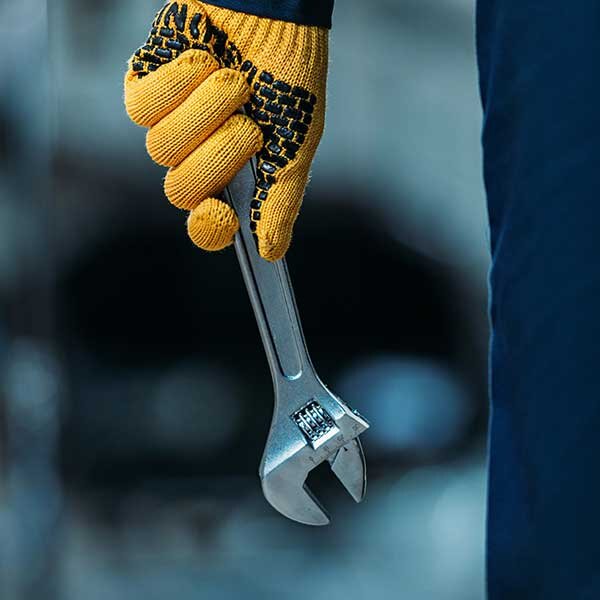 A hand wearing yellow gloves with blue palm grips holds a wrench by their side.