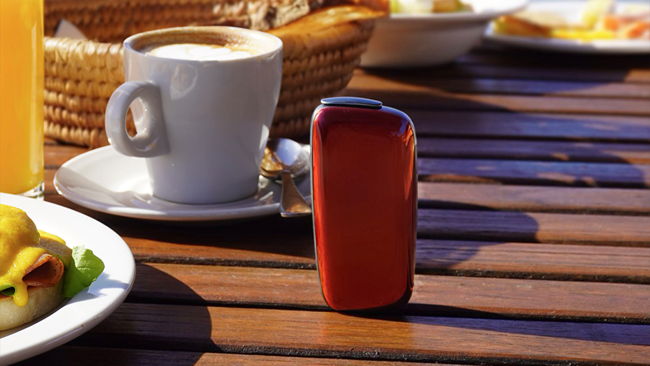 Dəniz mənzərəsi fonunda taxta masada qırmızı Ploom X Advanced cihazı və səhər yeməyi