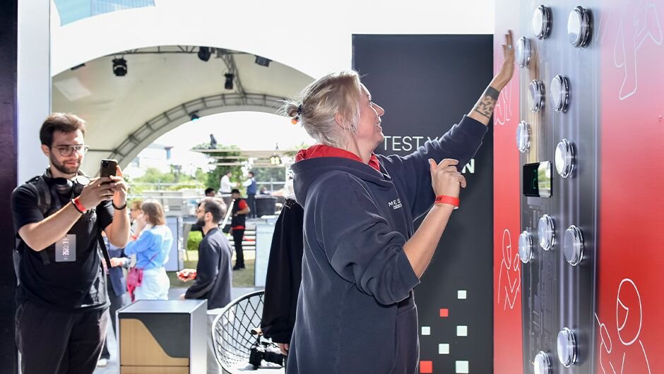 A female participant tests her reflexes in the Reaction Lab game at the Ploom Zone during Baku Race 2025, while other fans watch and take photos.