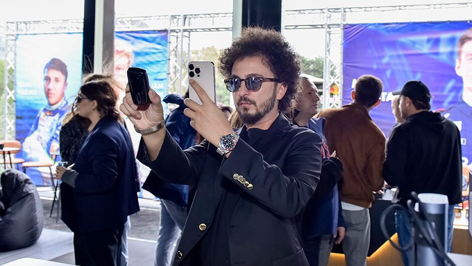 Kişi Ploom zonasında məhsulu və telefonunu çəkərək sosial mediada paylaşmaq üçün foto və video hazırlayır, arxa planda yarışçıların posterləri görünür 