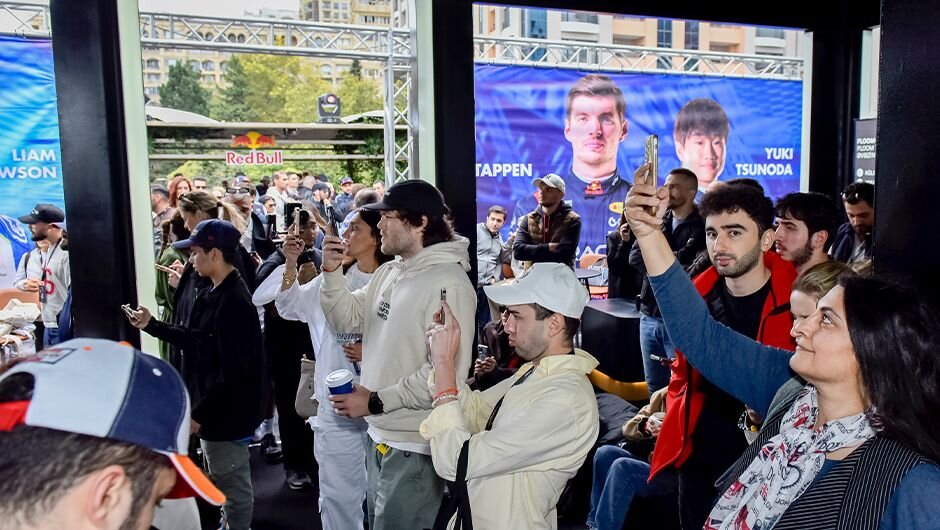 A man records the Ploom product and his phone in the Ploom Zone to share on social media, with race driver posters in the background.