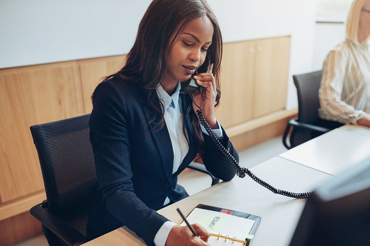 African American businesswoman writing down notes and talking on a telephone while working with a colleague at a desk in an office reception area