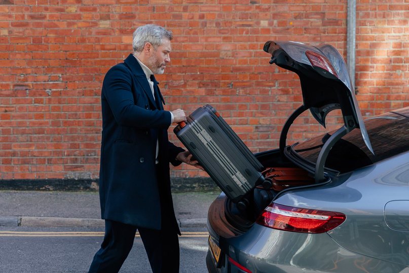 A man dressed in black places a grey suitcase into the boot of a grey sports car. 