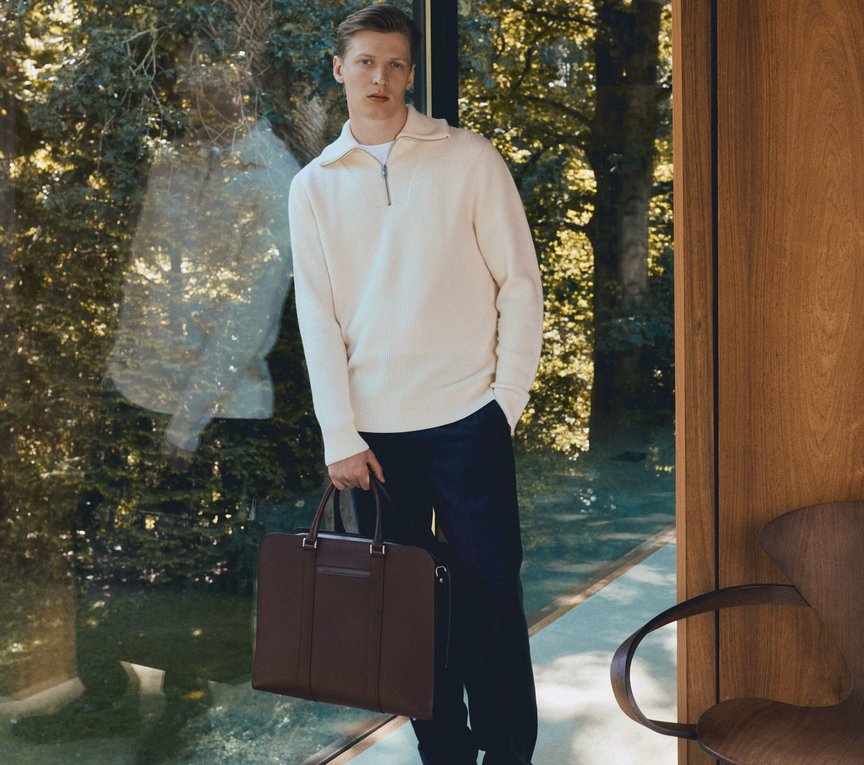 Man in white jumper and black trousers leans on a glass wall holding premium brown leather briefcase