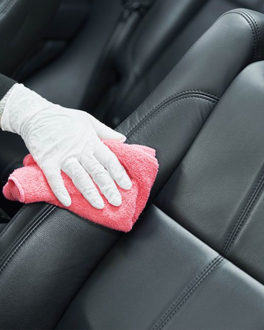 Gloved hand cleaning black PU leather car seat with pink microfibre cloth
