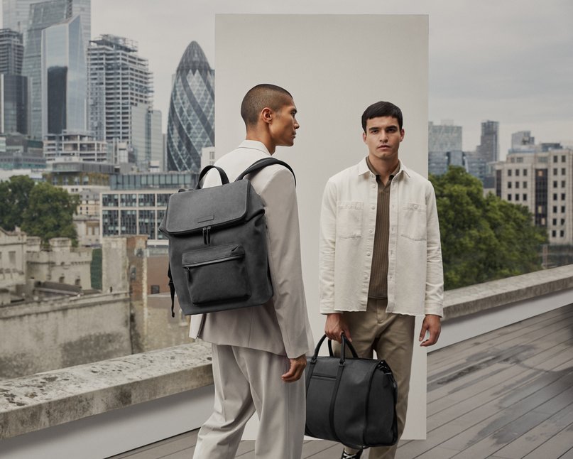 Two models stand holding grey backpack and weekend bag with London skyline behind