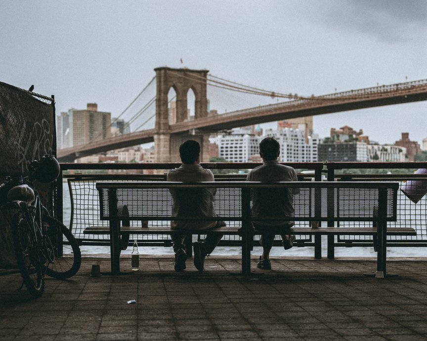 Two men sitting on a bench facing a large bridge with river flowing below