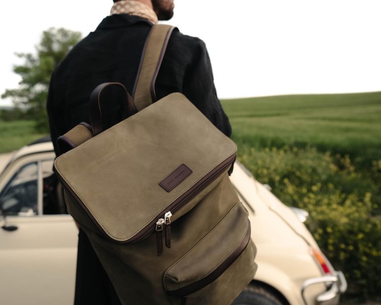 Man with green leather backpack on shoulder looks over green field with cream car in background