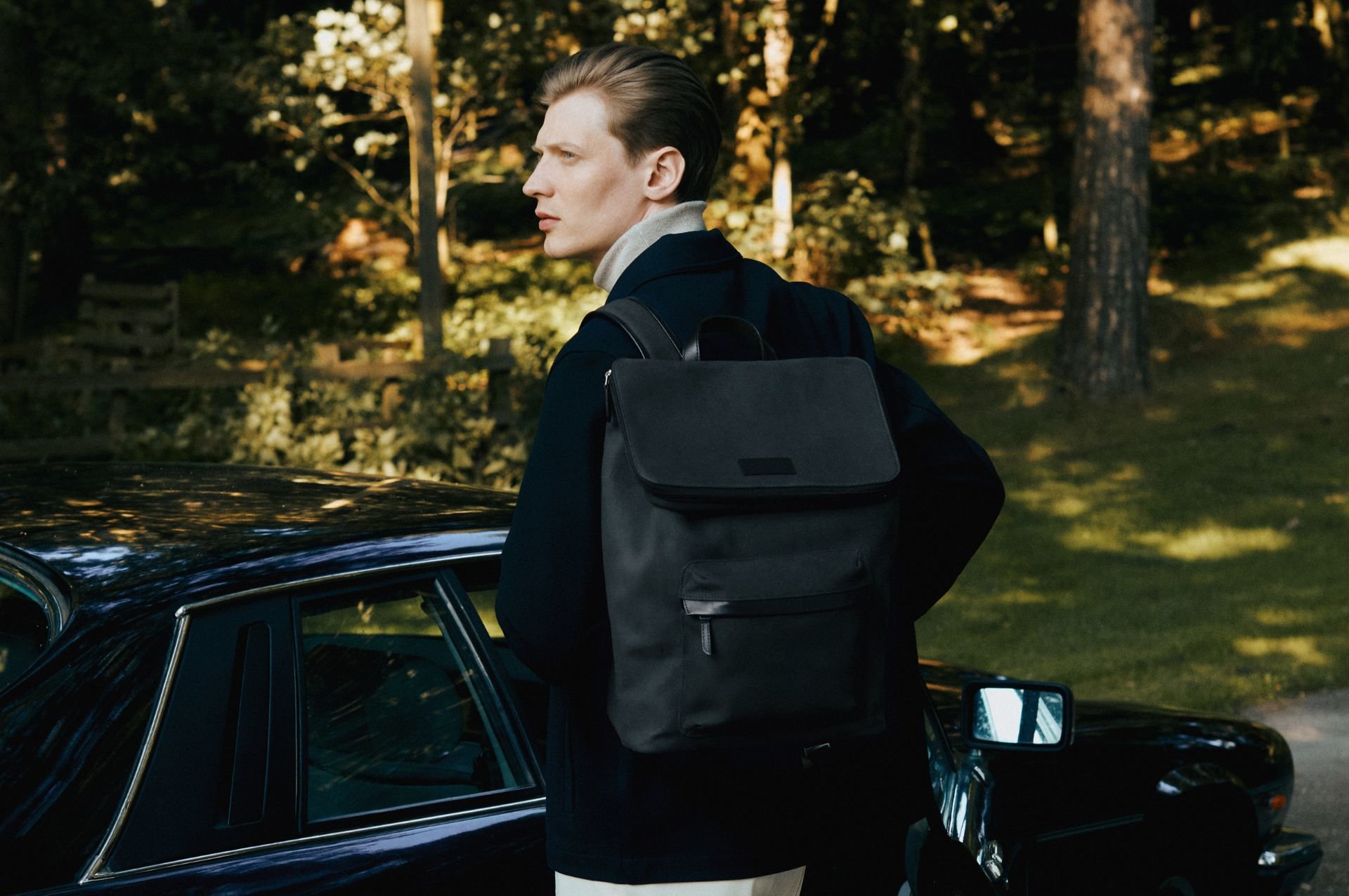 Man wearing blue coat and grey nubuck backpack looks back, with black car in background