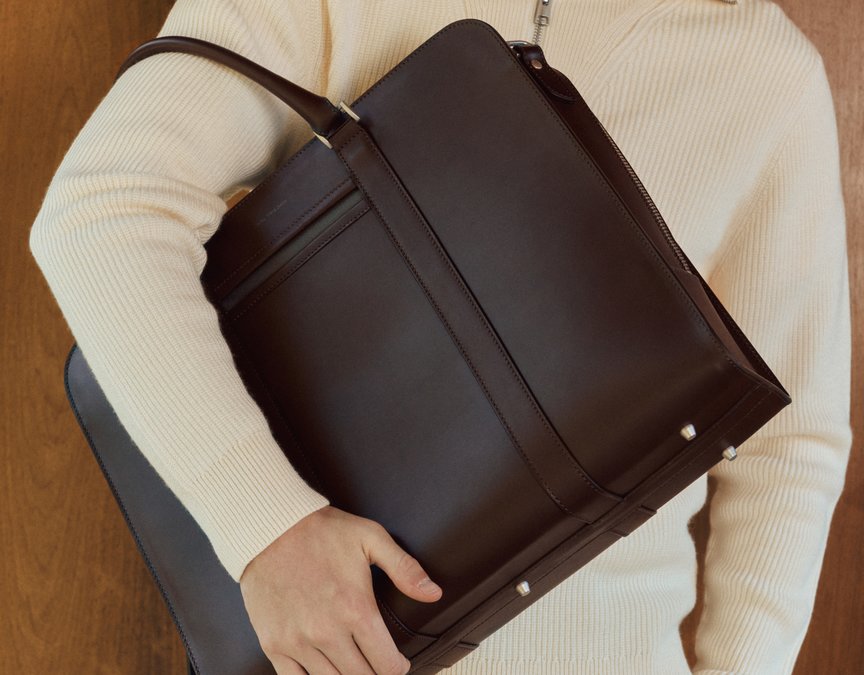 Man in white jumper holds premium brown leather briefcase over his upper arm