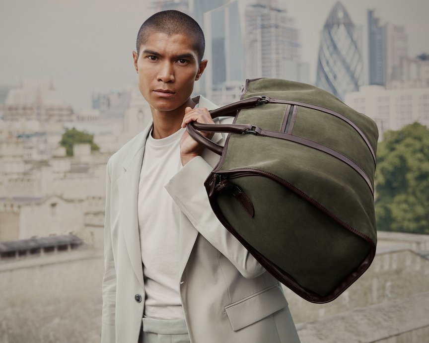 Man holds green weekend bag over shoulder with London skyline behind