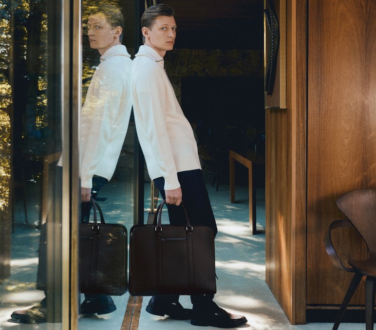 Man wearing long white jacket and brown large briefcase leans on glass wall which shows his reflection