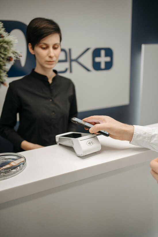 A smiling woman in a red sweater makes a payment by tapping her card on a contactless payment terminal inside a modern, well-lit business space.