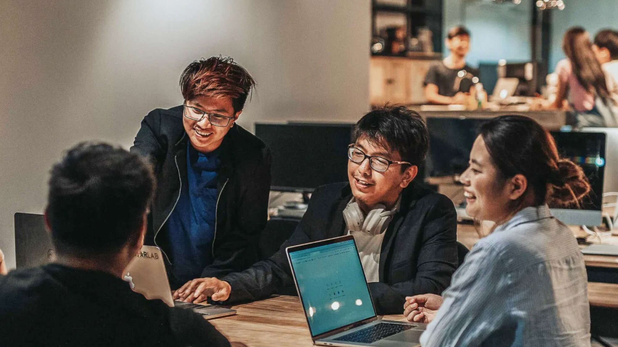 A diverse group of four business professionals in a modern office discussing strategy around a table, with laptops and documents visible, symbolizing collaboration between finance and engineering teams.