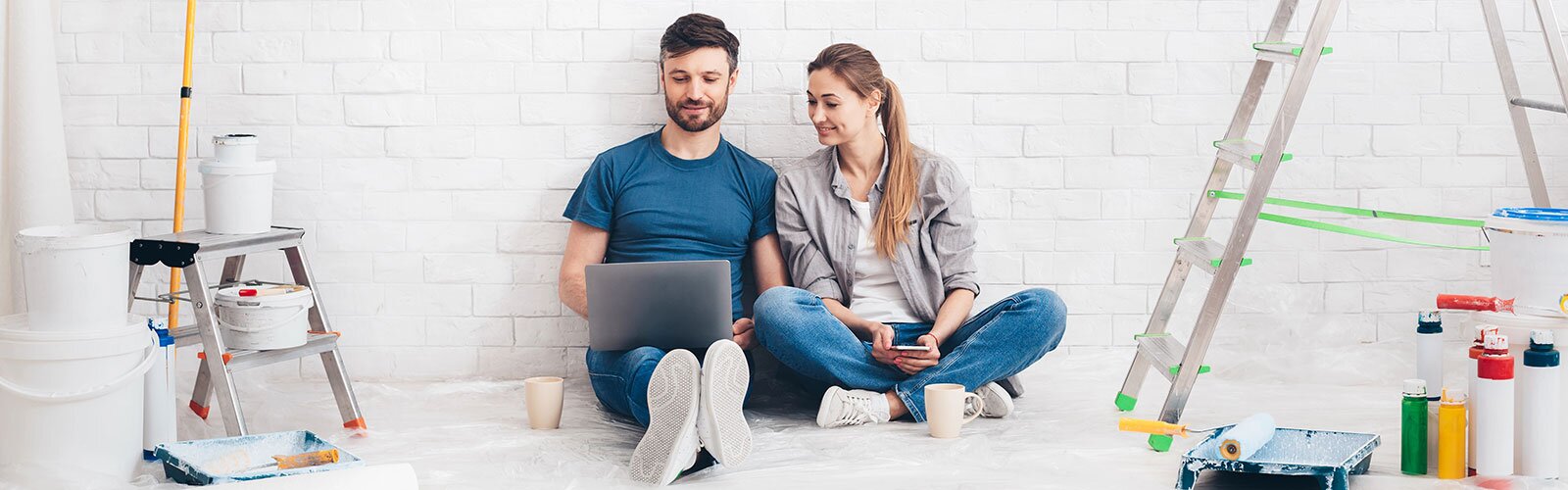 A couple pleasantly sits on the floor looking at their phones surrounded by painting supplies.