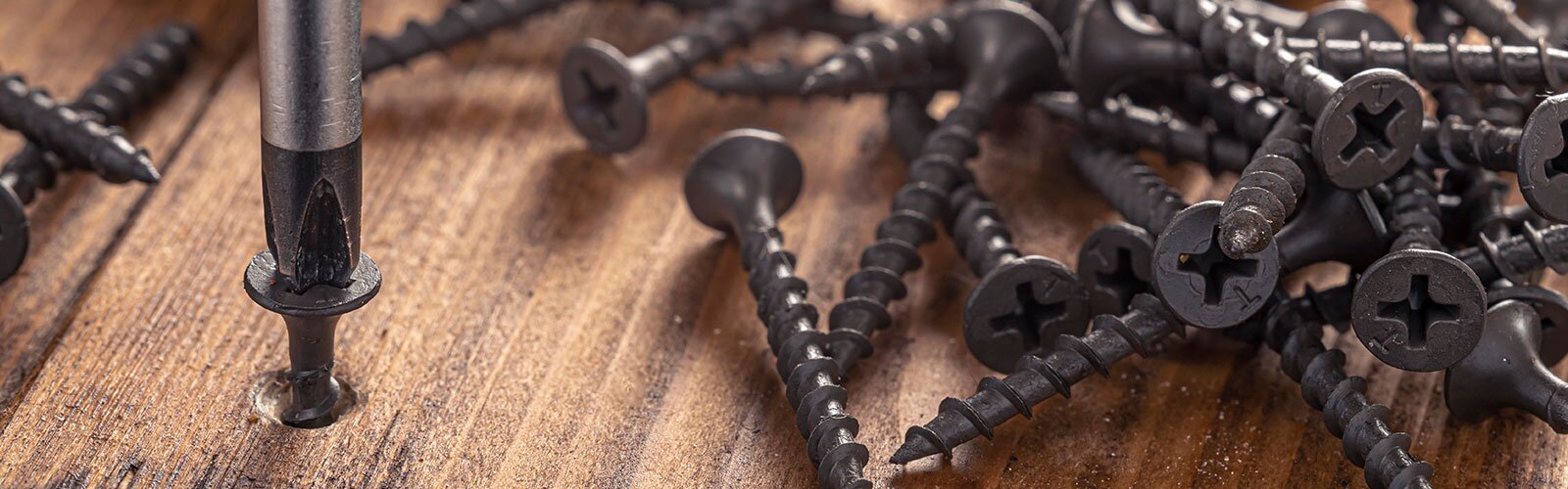 A pile of nails on a wooden surface, one being screwed into a wooden plank with a screwdriver.