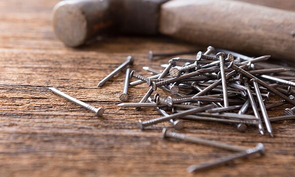 A pile of nails on a wooden surface, a hammer sits out of focus in the background.
