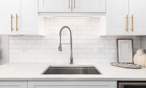 A white kitchen counter and cabinets with an undermount sink, faucet, and lighting.