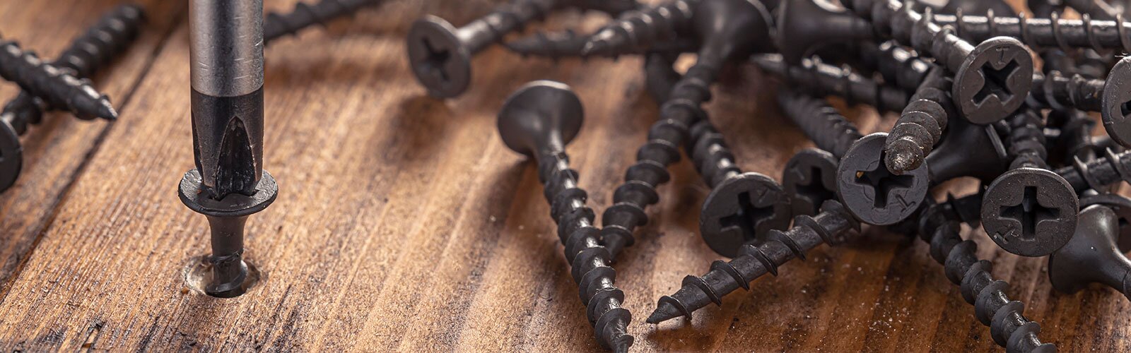A pile of nails on a wooden surface, a screwdriver screws in one of the nails.