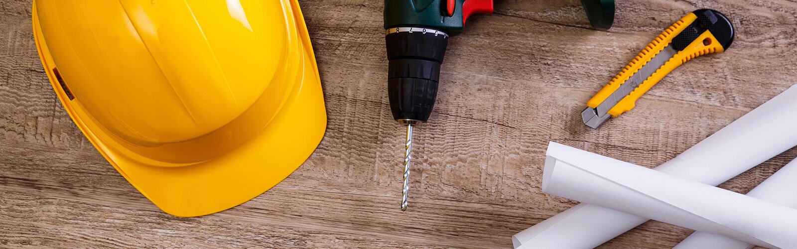 A yellow hard hat, a drill, blueprints, and an exacto knife lying on a wooden surface.