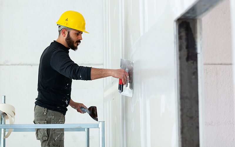 A contractor on scaffolding applies plaster to drywall, finishing a space.