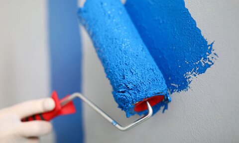 A hand holding a paint roller with blue paint begins to paint a white wall.