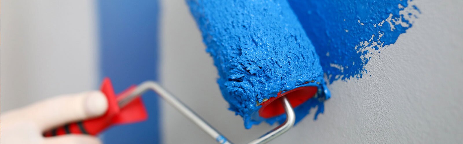 A hand holding a paint roller with blue paint begins to paint a white wall.