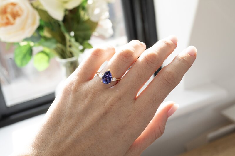 Model wearing an elegant yellow gold ring featuring a large, pear-shaped blue tanzanite gemstone.
