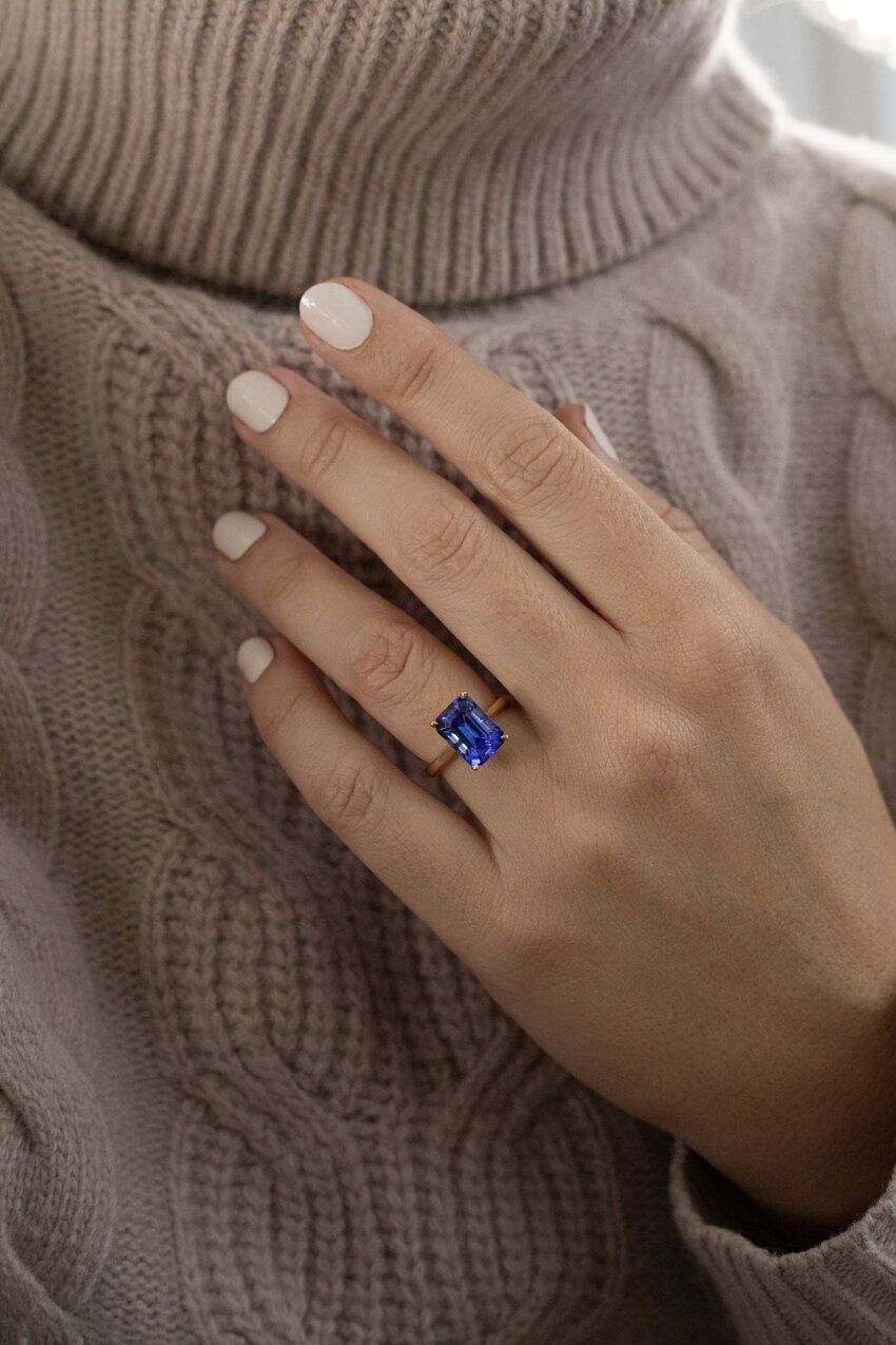 Model wearing a yellow gold solitaire ring with a large emerald-cut blue tanzanite gemstone.