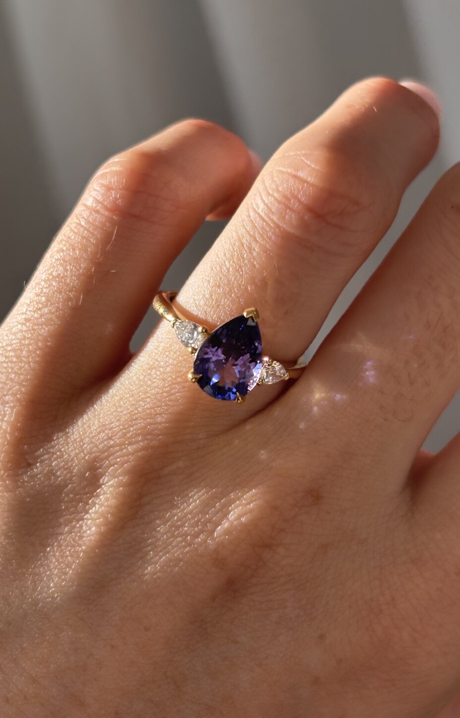 Model wearing a yellow gold ring set with a pear-shaped blue tanzanite and diamond side details.
