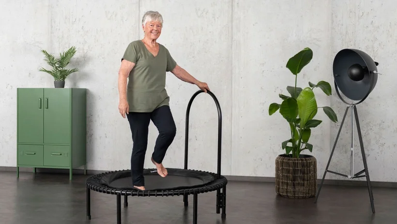 An older woman is working out on the bellicon rebounder, using the side handlebar for support.