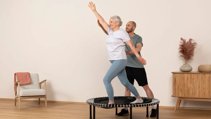 An older woman is accompanied by a physiotherapist during her rebounder workout.