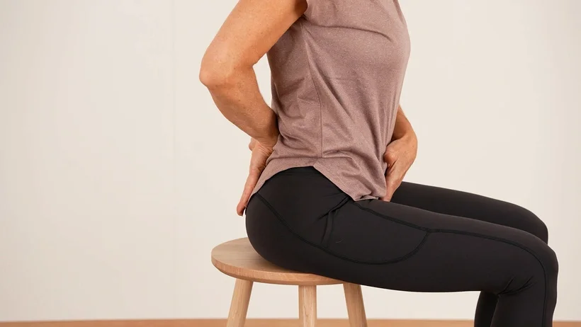 Jacqueline Huber sitzt auf einem Hocker und zeigt die optimale Beckenstellung mit ihren Händen.