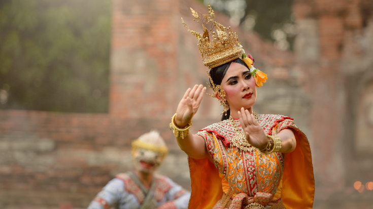 Een afbeelding van een Thaise vrouw in traditionele klederdracht - met een verfijnde gouden kroon op haar hoofd, dikke zwarte wenkbrauwen en zwart haar, en een jurk in verschillende tinten oranje met ingewikkelde gouden patronen, ter illustratie van een blogpost met de titel '33 leuke weetjes over Thailand'.