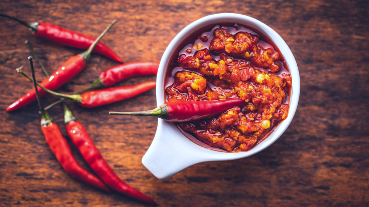 Een afbeelding van een witte kom op een houten tafel, gevuld met olie met daarin dunne Thaise chilipepers. De chilipepers en olie zijn rood en dit dient ter illustratie van een blogpost met de titel '33 leuke weetjes over Thailand'.