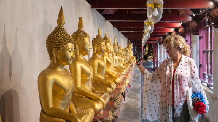 Een afbeelding van een vrouw van middelbare leeftijd met blond krullend haar en een shirt in kimonostijl die met haar mobiele telefoon een foto maakt van een rij gouden Boeddhabeelden in een tempel in Thailand, ter illustratie van een blogpost met de titel '33 leuke weetjes over Thailand'.
