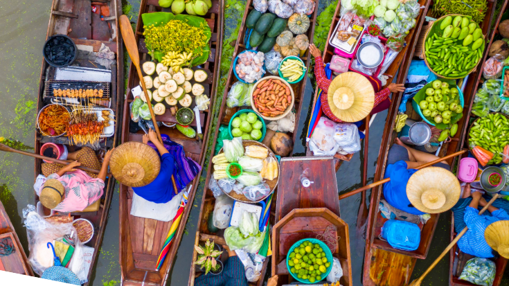 Een luchtfoto van vrouwen op traditionele houten Thaise longtailboten die op een rivier in Thailand verse producten verkopen, waaronder gele bananen, oranje papaja's, groene stekelige doerians en bereid eten zoals vlees op een stokje en garnalen. Ter illustratie van een blogpost met de titel '33 leuke weetjes over Thailand'.