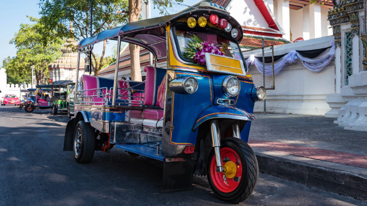 Een afbeelding van een felgekleurde tuk-tuk in Thailand, geparkeerd langs een stoeprand met een blauwe kap, roze zijkanten en gele, roze en blauwe lampen die de bovenkant van de voorkant sieren, ter illustratie van een blogpost met de titel '33 leuke dingen om te doen in Thailand'.