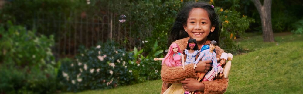 A girl holds a collection of Barbie dolls and a Ken doll, smiling while running through a yard.