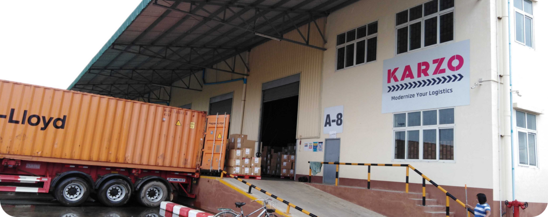 A large warehouse loading dock with an orange shipping container (Maersk-Lloyd) backed up to the bay, surrounded by boxes in front of Karzo Building