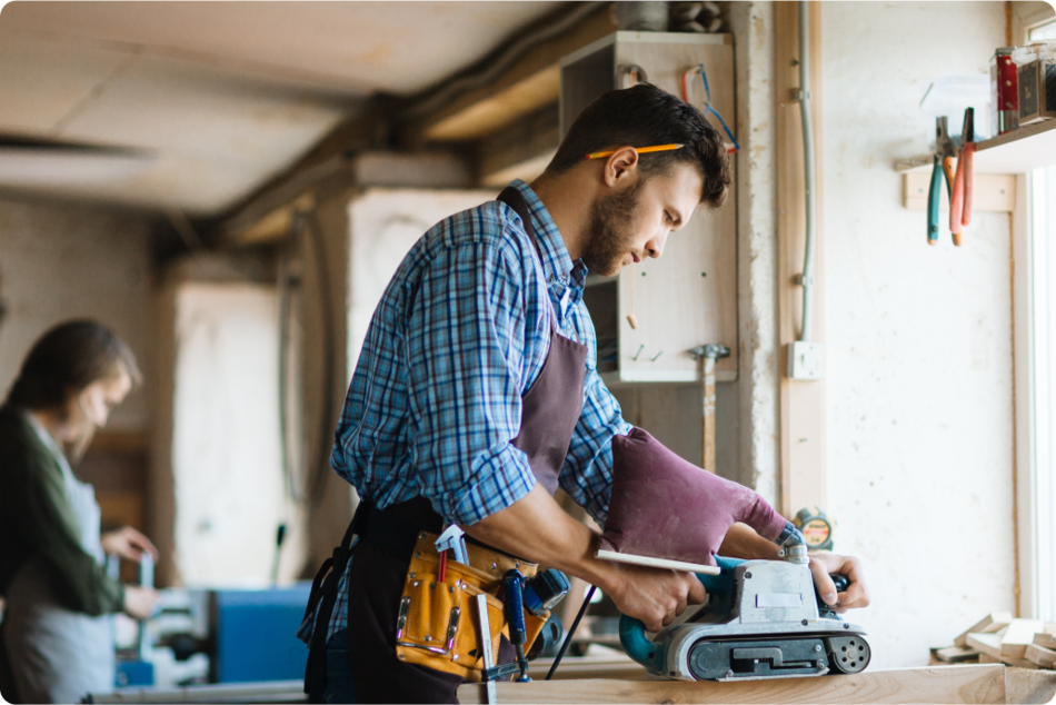 A man with pencil in his ear, wearing a construction toolbelt and operating a power sander.